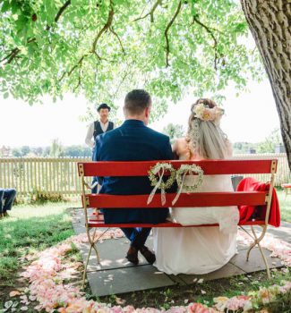 Brautpaar auf einer roten Bank unter Apfelbaum während der freien Trauung