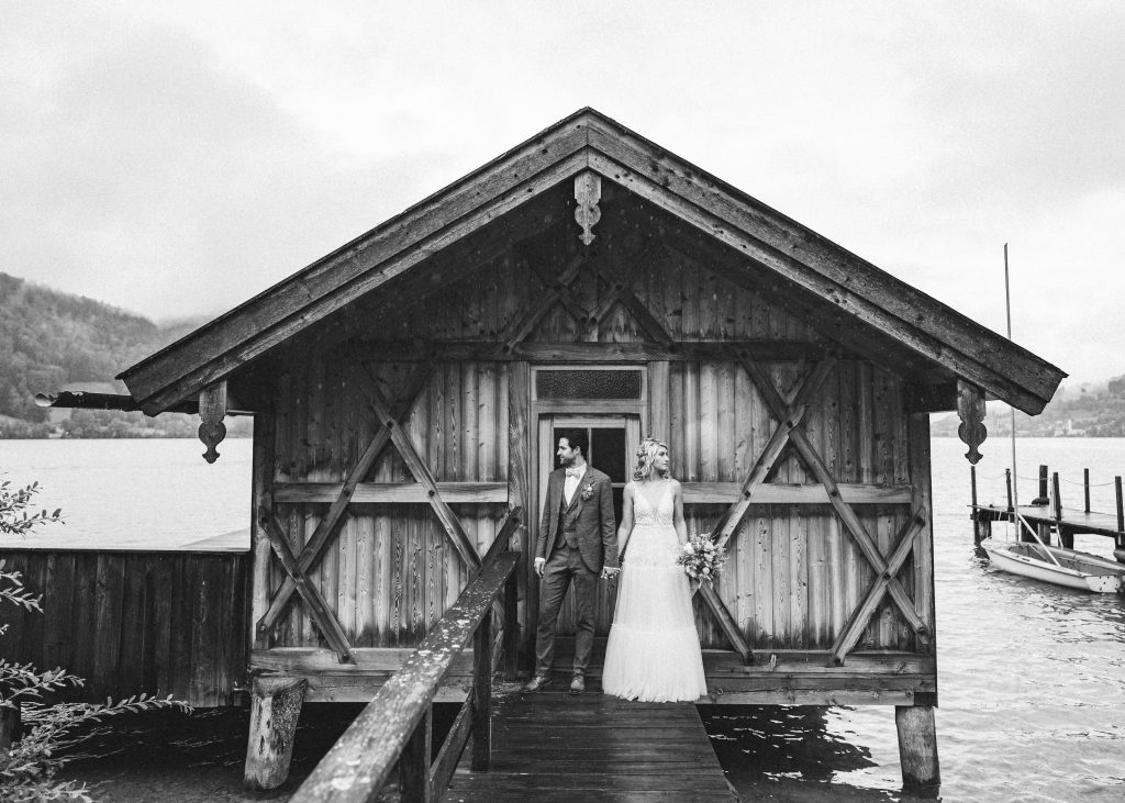 Hochzeitsfotograf München: Alex Mayer | Brautpaar vor Holzhütte auf dem Steg am See | Strauß & Fliege
